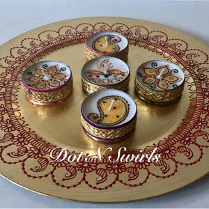 May include: A gold-colored decorative plate with a red henna-style border and five small, round candles. The candles have intricate designs and gold-colored rims. The text "Dot N Swirls" is visible on the plate.