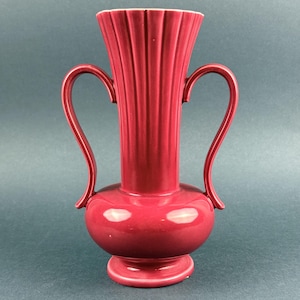 May include: A red ceramic vase with two curved handles. The vase has a bulbous base and a fluted, cylindrical neck. The vase is a deep red color and sits on a small, round base. The handles curve gracefully from the neck to the body.
