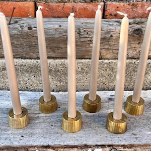 May include: Six tall, slender, light beige taper candles, each standing upright in a circular, brushed gold-toned metal candle holder with a slightly tapered base. The candles are arranged in two rows of three on a weathered, light gray wooden surface.