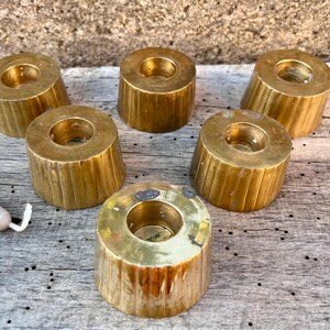 May include: A set of six cylindrical brass candle holders with vertical fluted detailing along the sides, each featuring a central circular opening for a taper candle, accompanied by two light gray taper candles with white wicks.