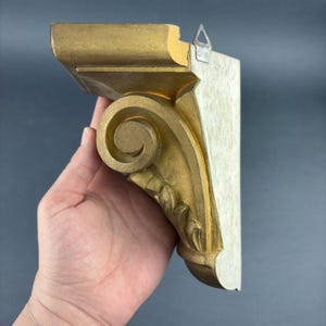 Op de afbeelding: Een goudgeverfde houten kraagsteen met een sierlijk rolontwerp. De kraagsteen heeft een platte bovenkant en een haakse vorm, met een metalen hanger aan de achterkant. De kraagsteen wordt in een hand gehouden.