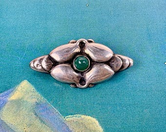 May include: A silver-toned metal brooch featuring a symmetrical, elongated floral-inspired design with smooth, rounded petals radiating from a central, circular, polished green stone cabochon set firmly in the middle of the piece.
