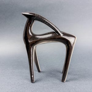 Può includere: Una scultura in ceramica marrone scuro lucida di un cervo stilizzato. La scultura ha un design elegante e moderno con gambe lunghe e sottili e una schiena curva. Il collo del cervo presenta un motivo inciso decorativo.