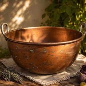 Peut inclure: Un grand saladier en cuivre antique avec deux poignées incurvées. Le bol a un design large et ouvert et une patine vieillie. Il repose sur une surface en bois avec un tissu blanc, des raisins, des figues et des herbes. Le fond présente un feuillage vert.