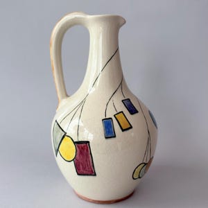 May include: A cream-coloured ceramic pitcher with a crackle glaze finish. The body features hand-painted black lines resembling hanging strings, decorated with colourful geometric shapes including yellow circles, red rectangles, blue squares, and orange accents.