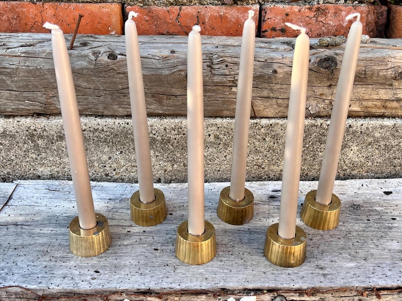May include: Six tall, slender, light beige taper candles, each standing upright in a circular, brushed gold-toned metal candle holder with a slightly tapered base. The candles are arranged in two rows of three on a weathered, light gray wooden surface.
