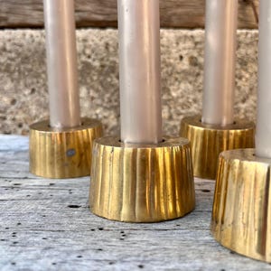 May include: A set of four brass candle holders with a fluted, tapered base design. Each holder features a smooth, cylindrical top section designed to securely hold a single light-colored taper candle in an upright position.