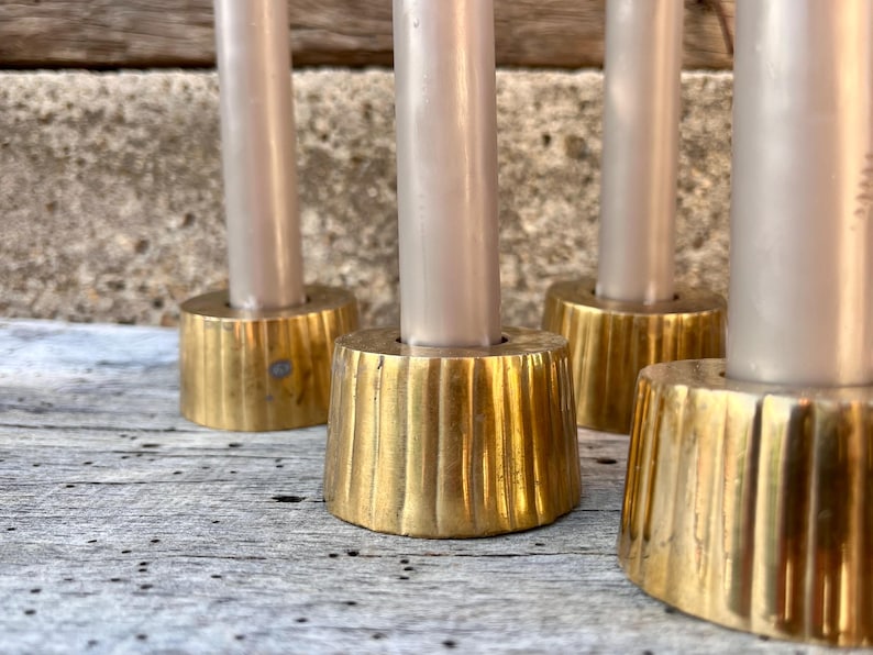 May include: A set of four brass candle holders with a fluted, tapered base design. Each holder features a smooth, cylindrical top section designed to securely hold a single light-colored taper candle in an upright position.