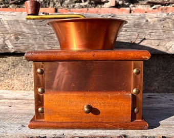 May include: A manual coffee grinder featuring a square wooden base with a small pull-out drawer and a central copper panel secured by four brass rivets. A large copper hopper sits on top, equipped with a gold-toned metal handle and a brown knob.