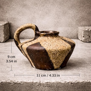 Puede incluir: Un jarro de cerámica vintage con asa. El jarro es marrón y beige con un patrón geométrico. Mide 9 cm de alto y 11 cm de ancho.
