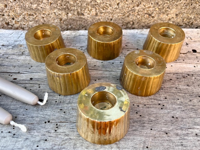 May include: A set of six cylindrical brass candle holders with vertical fluted detailing along the sides, each featuring a central circular opening for a taper candle, accompanied by two light gray taper candles with white wicks.