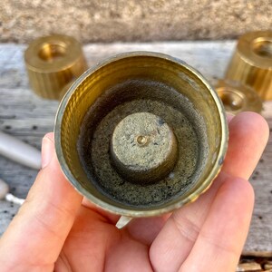 May include: A circular brass candle holder with a central spike for securing a candle. The interior features a textured, weathered finish, while the exterior has a ribbed design. Additional matching brass candle holders are visible in the background.