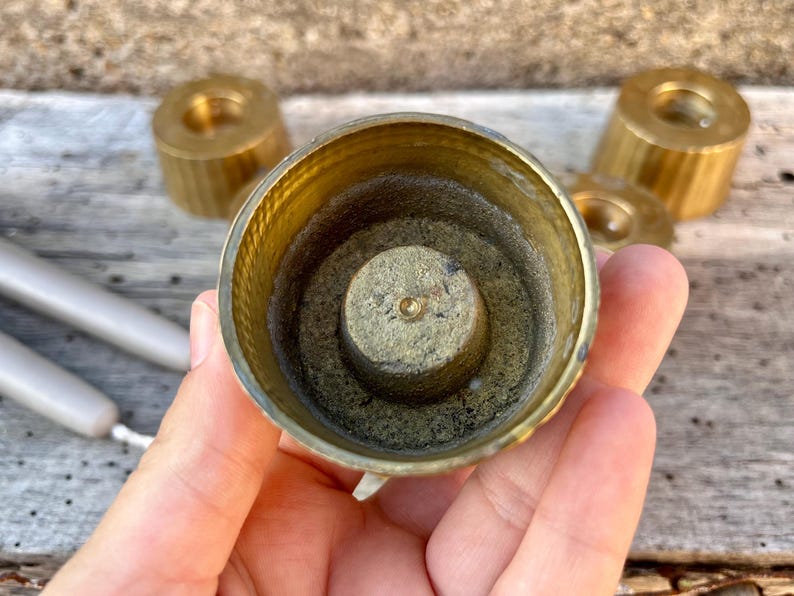 May include: A circular brass candle holder with a central spike for securing a candle. The interior features a textured, weathered finish, while the exterior has a ribbed design. Additional matching brass candle holders are visible in the background.