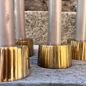 May include: A set of four cylindrical brass candle holders with a vertical ribbed texture, each holding a smooth, light gray taper candle. The metallic bases have a polished gold-tone finish and a wide, stable circular footprint for support.