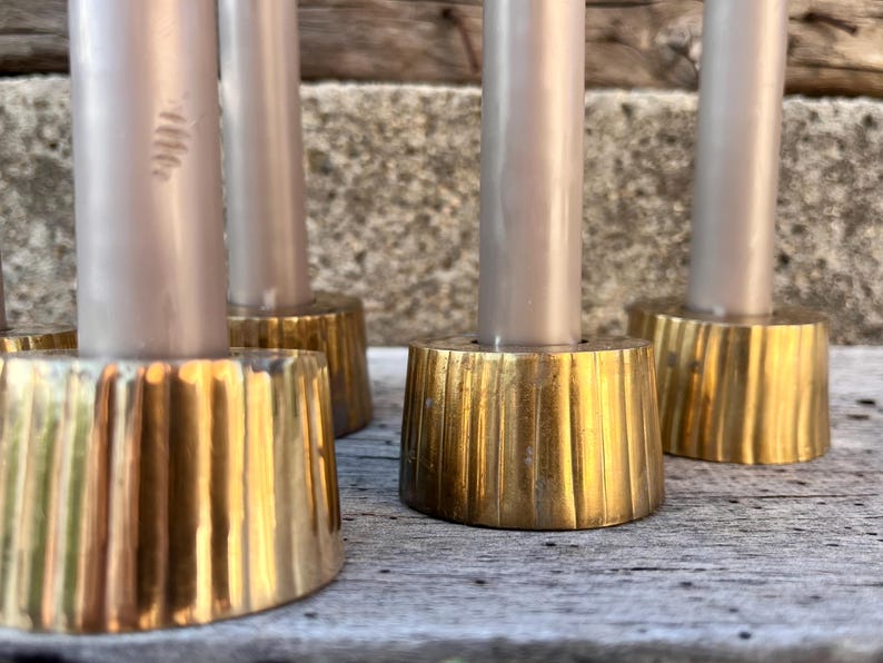 May include: A set of four cylindrical brass candle holders with a vertical ribbed texture, each holding a smooth, light gray taper candle. The metallic bases have a polished gold-tone finish and a wide, stable circular footprint for support.