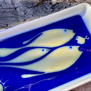 Könnte beinhalten: Rechteckige Servierplatte aus Keramik mit weißem Rand und blauem Innenraum. Die blaue Oberfläche zeigt ein Design von drei gelben Fischen mit blauen Augen. Ideal zum Servieren von Vorspeisen oder zum Präsentieren von Dekorationsartikeln.