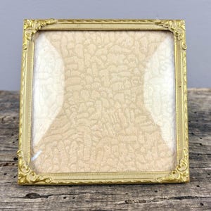 Op de afbeelding: Vierkante, antiek goudkleurige fotolijst met sierlijke hoekdetails. De lijst heeft een glazen voorkant en een crèmekleurige achterkant met patroon. De lijst staat op een verweerd houten oppervlak.
