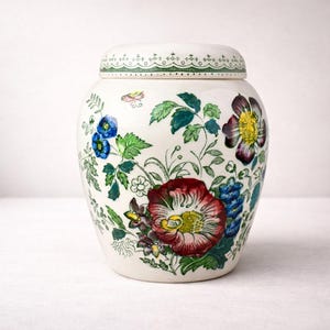 Op de afbeelding: Een crèmekleurige keramische pot met bijpassend deksel, voorzien van een kleurrijke botanische print van rode, blauwe en paarse bloemen met groene bladeren. Het deksel heeft een decoratief groen geschulpt randpatroon langs de bovenrand.
