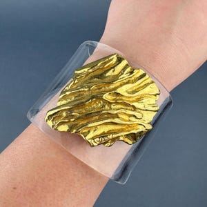 May include: A wide cuff bracelet featuring a clear, transparent acrylic band. Centered on the band is a large, irregular, textured gold-tone metal piece with a molten, organic surface finish that covers a significant portion of the clear cuff.