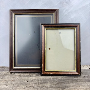 Peut inclure: Deux cadres photo rectangulaires avec des bordures en bois brun foncé et une fine garniture intérieure dorée. Le plus grand cadre à gauche contient un fond gris foncé, tandis que le plus petit à droite présente un fond beige clair et une vitre.