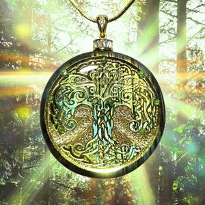 May include: A gold-colored pendant necklace featuring a detailed tree of life design. The pendant has a circular shape with intricate patterns and small green accents. The necklace hangs against a blurred forest background.