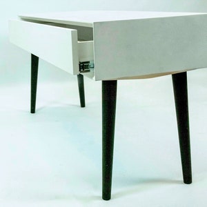 May include: White side table with a single drawer and black tapered legs. The drawer is open and the interior is white.