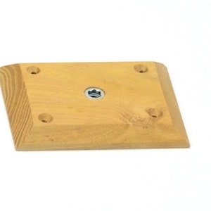 May include: A light brown wooden climbing hold with four screw holes and a central mounting hole. The hold has a slightly rounded, rectangular shape.