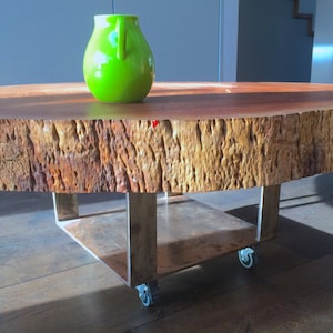 May include: A round wooden coffee table with a natural wood grain finish and a metal base with wheels. A green ceramic vase sits on the table.