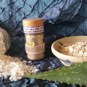 May include: A small glass jar with a wooden lid and a colorful patterned label that reads "Rice Wash". The jar is filled with a light brown powder. The image also shows a bowl of rolled oats, a pile of white rice, and a green aloe vera leaf.