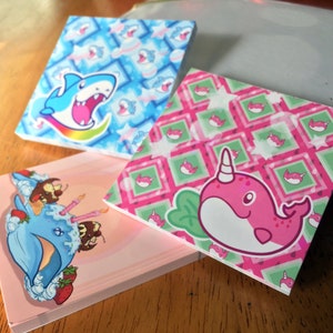 May include: Three small pink, blue and green paper notepads with different animal designs. The pink notepad has a pink narwhal with a green leaf and a white background. The blue notepad has a blue shark with a rainbow tail and a white background. The green notepad has a pink narwhal with a green leaf and a white background.