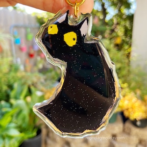Puede incluir: Llavero de gato negro con ojos amarillos, hecho de resina transparente con purpurina. El llavero tiene forma de gato sentado y mirando hacia un lado.