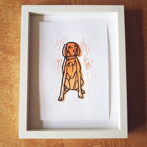 May include: A framed print featuring a stylized orange dog illustration with black outlines. The dog is sitting, and the print has an orange textured background. The white frame contrasts with the dog print and the wooden surface.