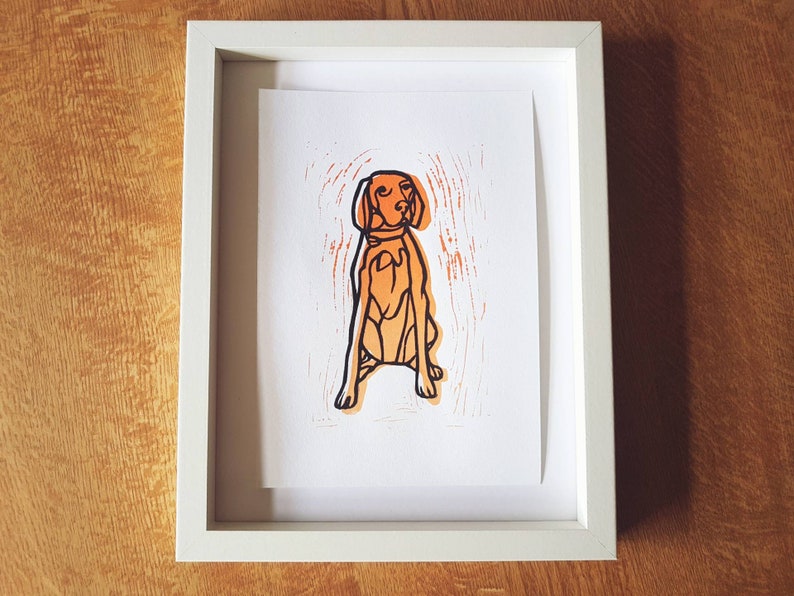 May include: A framed print featuring a stylized orange dog illustration with black outlines. The dog is sitting, and the print has an orange textured background. The white frame contrasts with the dog print and the wooden surface.
