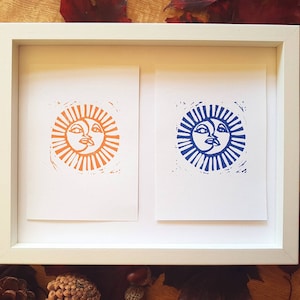 May include: Two framed prints of a sun and moon design. The print on the left is orange and the print on the right is blue.