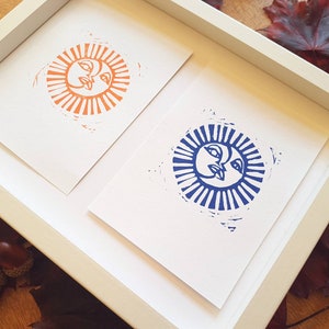 May include: Two framed prints of sun faces, one in orange and one in blue, with a white background.