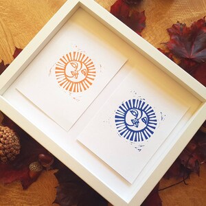 May include: Two framed prints of stylized sun faces. The sun on the left is orange with a white background. The sun on the right is blue with a white background.