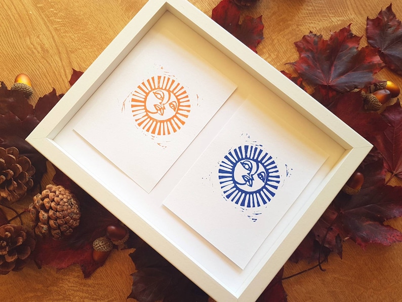 May include: Two framed prints of stylized sun faces. The sun on the left is orange with a white background. The sun on the right is blue with a white background.