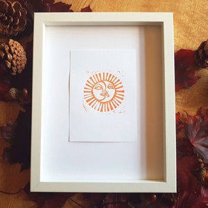 May include: A framed print of an orange sun with a face, set against a white background. The frame is white and has a simple design.