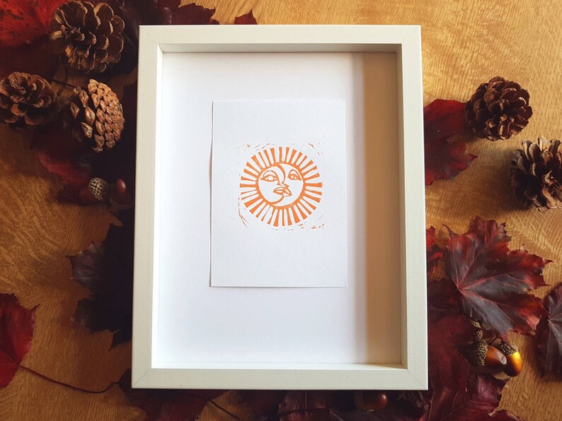 May include: A framed print of an orange sun with a face, set against a white background. The frame is white and has a simple design.