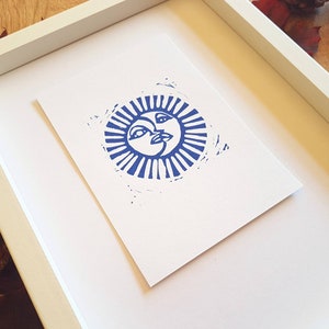 May include: A framed print of a blue sun with a face, surrounded by rays. The print is on white paper and is framed in a white frame.