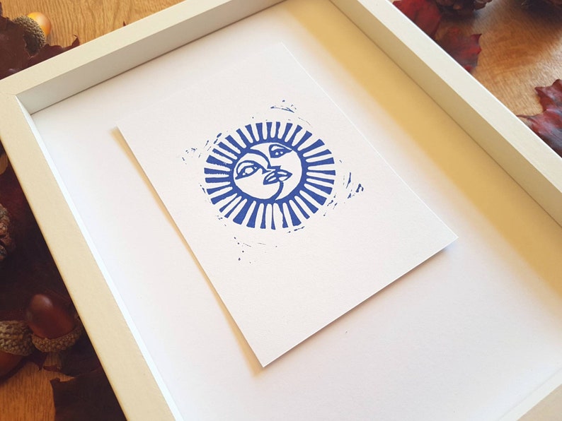 May include: A framed print of a blue sun with a face, surrounded by rays. The print is on white paper and is framed in a white frame.