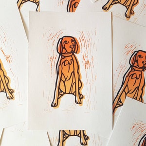 May include: A collection of art prints featuring a stylized orange dog illustration. The dog is depicted in a seated position with a collar, set against a white background. The prints have a woodcut or linocut style.