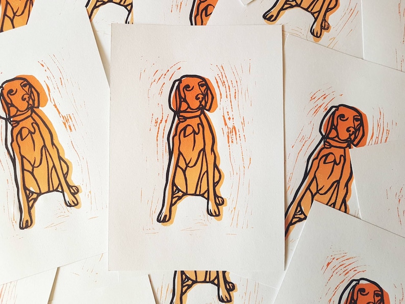 May include: A collection of art prints featuring a stylized orange dog illustration. The dog is depicted in a seated position with a collar, set against a white background. The prints have a woodcut or linocut style.