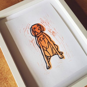 May include: A framed art print featuring a stylized orange and black dog illustration. The dog is depicted in a seated position with a collar, set against an orange textured background. The print is encased in a white frame.
