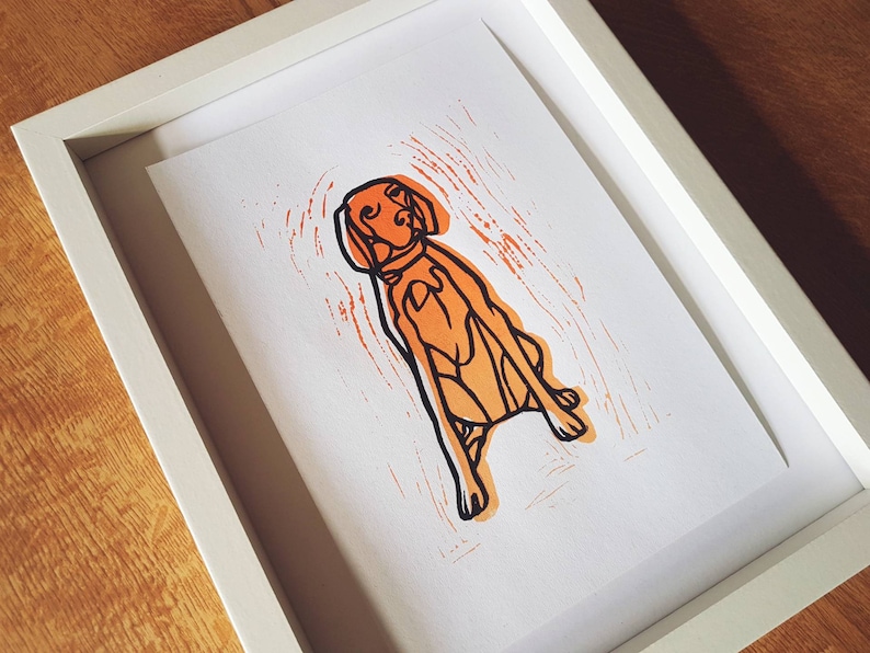May include: A framed art print featuring a stylized orange and black dog illustration. The dog is depicted in a seated position with a collar, set against an orange textured background. The print is encased in a white frame.