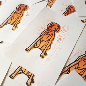 May include: A collection of art prints featuring a stylized orange and black illustration of a dog in a sitting position. The prints have a textured, woodcut-like appearance, with the dog's outline and details in black.