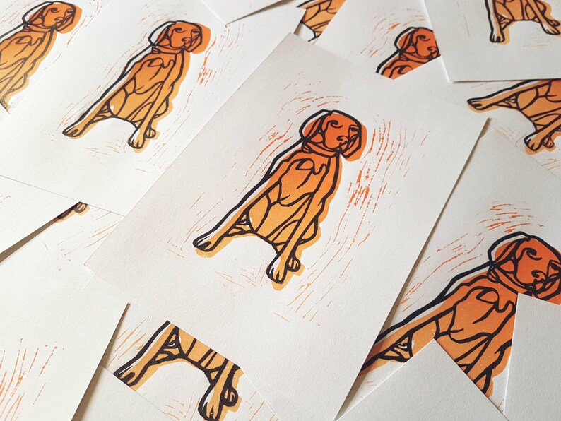 May include: A collection of art prints featuring a stylized orange and black illustration of a dog in a sitting position. The prints have a textured, woodcut-like appearance, with the dog's outline and details in black.