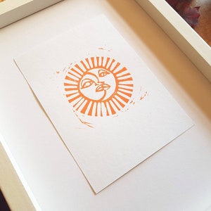 May include: A framed print of an orange sun with a face, surrounded by rays. The print is on white paper and is framed in a white frame.