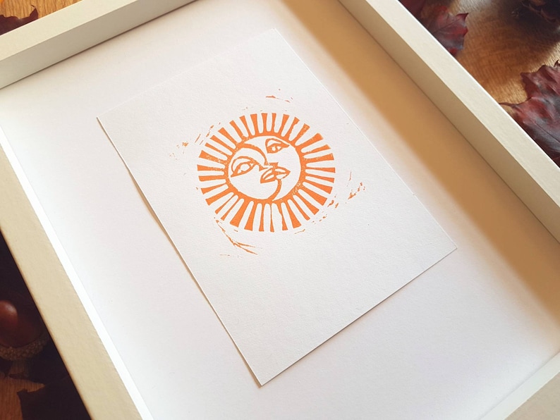 May include: A framed print of an orange sun with a face, surrounded by rays. The print is on white paper and is framed in a white frame.