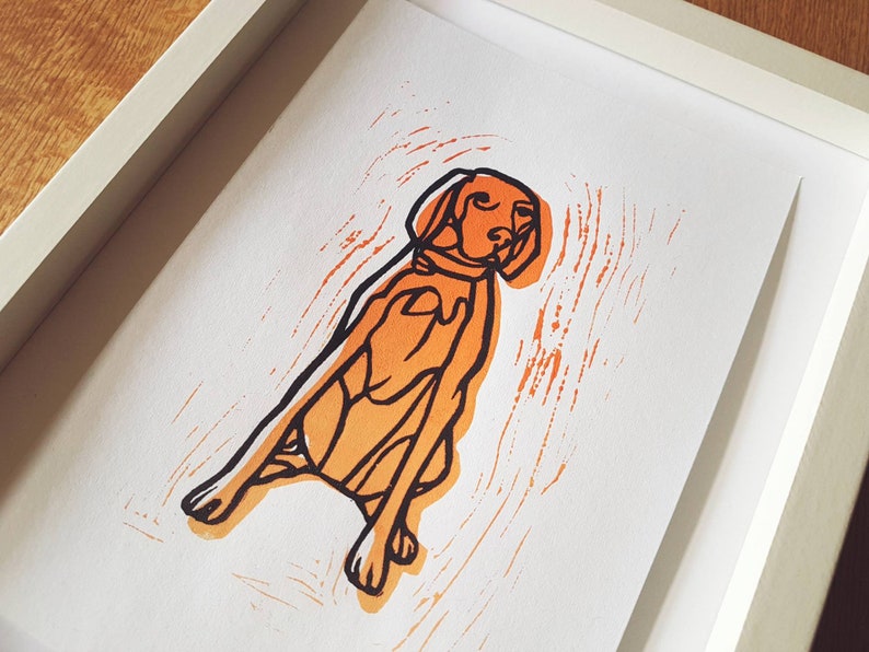 May include: A framed print featuring a stylized orange and black illustration of a dog. The dog is depicted in a seated position with a collar. The artwork is set against a white background within a white frame.